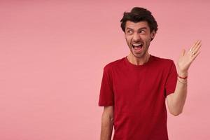 Furious aggressive young man with stubble in red t shirt arguing and screaming over pink background Looking to the side photo