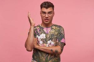 Attractive young man with trendy haircut wearing glasses and flowered t-shirt raising his hand and making indignant face, frowning crossly, isolated over pink background photo