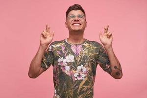 Indoor shot of happy guy with short haircut, dressed in casual clothes, gesturing with fingers, smiling broadly with eyes closed, expecting good news, isolated over pink background photo