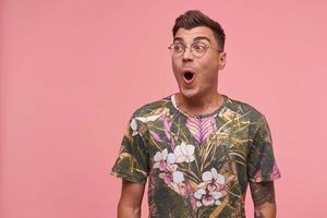 Studio portrait of handsome young lovely open-eyed male, looking aside with surprised face, puckering and folding lips, posing over pink background photo