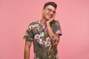 Indoor portrait of cute shy young guy with palm on his cheek, wearing glasses and flowered t-shirt, looking aside with blushing smile, standing over pink background photo