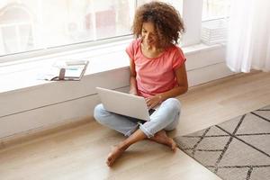 Young pretty dark skinned curly woman working remotely from home, sitting on floor near window with laptop on legs, looking at screen and smiling slightly photo