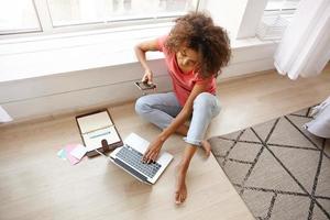 Top view of pretty dark skinned curly lady in casual clothes posing over home interior, working out of office with laptop and mobile phone, typing text on keyboard