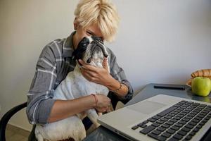 Indoor photo of young pretty woman with blond hair, wearing casual clothes, taking break while working home, making fun with her cute black and white dog