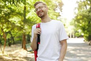 Outdoor portrait of young pretty bearded guy, looking ahead and smiling, wearing glasses and casual clothes, walking through park on sunny day photo