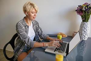 retrato de una mujer bastante joven e independiente con el pelo rubio corto, sentada al lado de una computadora portátil, comunicándose con alguien, de buen humor foto