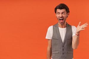 Angry irritated young man with stubble screaming and looking to the side isolated over orange background photo
