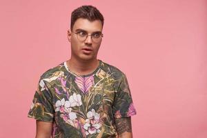 Indoor shot of open-eyed handsome short haired guy in casual t-shirt looking aside with confused face, posing over pink background with hands down photo