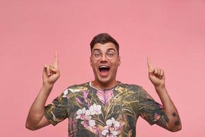 Indoor shot of happy attractive short haired man in glasses looking to camera with raised hands, contracting forehead and smiling broadly to camera, isolated over pink background photo