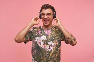 Indoor shot of beautiful young man wearing glasses and flowered t-shirt standing over pink background with raised to ears hands, enjoying music in his headphones, looking happy and pleased photo