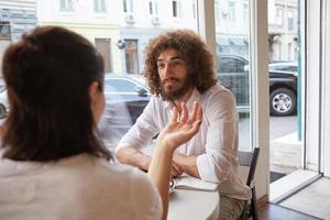 retrato interior de un hermoso hombre rizado con barba que se reunió en un café, mirando con atención y calma a la mujer a su lado, sentado en la mesa junto a la ventana foto