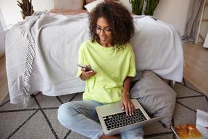 Pleasant looking young dark haired female with curls working on floor of bedroom, holding smart phone in hand and keeping laptop on legs, receiving good news, smiling joyfully photo