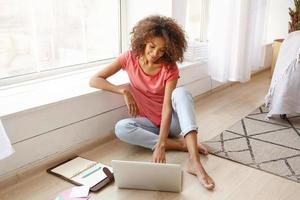 Indoor shot of attractive dark skinned woman leaning on windowsill while sitting on floor, working remotely from home with modern laptop and notebook, positive emotions