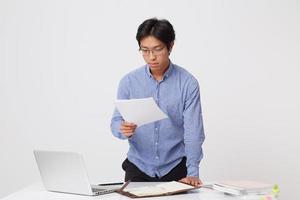 Concentrated asian young business man in glasses using laptop and earphones for video communication working with documents over white background photo