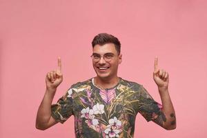 Happy young dark haired man in casual t-shirt smiling to camera with raised hands, pointing with fingers upwards, standing over pink background photo