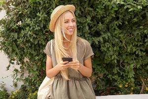 Outdoor portrait of cute young blond female in casual linen dress over green garden, keeping smart phone in hands and looking aside with broad smile photo
