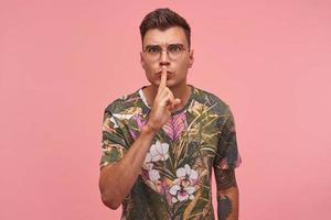 Close up of attractive young man with tattoos keeping index finger on lips, asking to keep secret, do not make noise, demonstrating silence gesture, standing over pink background photo