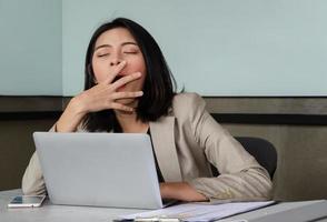 joven mujer de negocios bostezando en la mesa de la oficina frente a la computadora portátil, cubriendo su boca por cortesía. concepto de exceso de trabajo y privación del sueño foto