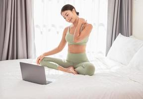 Woman practicing yoga on bed after waking up. Asian female sitting on bed using her laptop while doing stretching exercise at home.