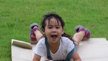 una niña sonriente se sienta en una caja de cartón deslizándose por una colina en un jardín botánico. el famoso centro de aprendizaje al aire libre de mae moh mine park, lampang, tailandia. concepto de infancia feliz. video