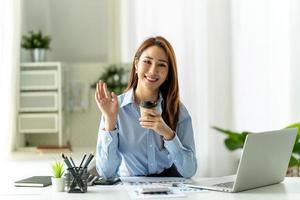 Asian woman sitting at wooden desk in office, looking at camera, waving hello. photo