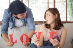 Beautiful young asian couple sitting on sofa holding word love together in living room, family husband and wife smiling with happy, man and woman lover wearing casual with relax, lifestyle concept. photo