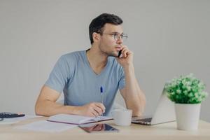 Serious male finance manager talks on mobile phone, solves problems with partner, writes some notes in diary, arrange meeting, surrounded with laptop computer, isolated over grey background. photo
