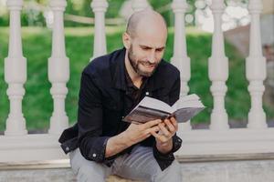 Close up shot of focused attractive bald young man holds book, reads with joy, enjoys weekends, spends free time in summer garden, likes reading in calm atmosphere. People and relax concept. photo