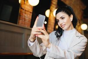 Beautiful woman with dark hair tied in pony tail making selfie while sitting in cafe using her smartphone. Well-groomed female in elegant clothes looking in screen of her telephone making photo