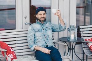 Photo of pleased blue eyed bearded male dressed in denim clothing, raises hand with cup of coffee or tea, being in good mood, rests in outdoor cafeteria with cozy interior and calm atmosphere