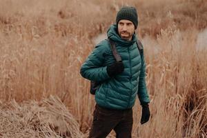 People and journey concept. Shot of handsome man dressed in warm jacket and headgear, walks outside, looks attentively aside, poses against field background with copy space on left side for text photo