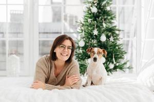 Joyful woman with short dark hair wears spectacles and round glasses, funny dog in spectacles poses near host, lie on bed, have good rest during Christmas eve, have fun together. New Year decoration photo