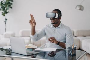 Excited afro man in vr glasses at workplace at home. Businessman has virtual meeting. photo
