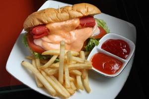 Complete hotdog package. Red sausage, cheese sauce, tomato wedges, lettuce. Plus french fries and a container of red chili and tomato sauce. Served on a white square plate. Close up. top view photo