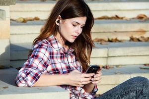 Image of beautiful stylish woman holding mobile phone. Young european girl standing at the street and using cellphone. Woman listening to music with the phone and having fun. Mobile internet concept. photo