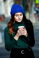 joven elegante bebiendo té en una calle de la ciudad. hipster chica europea con vaso de papel con leche. hermosa mujer joven con una taza de café en la calle de la ciudad. descanso. Café para llevar. foto