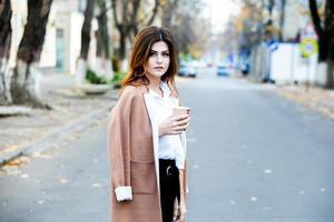 joven elegante bebiendo té en una calle de la ciudad. hipster chica europea con vaso de papel con leche. hermosa mujer joven con una taza de café en la calle de la ciudad. descanso. Café para llevar. foto