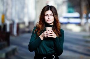 Young stylish woman drinking tea in a city street. Hipster european girl with latte paper glass. Gorgeous young woman with cup of coffee in city street. Coffee break. Coffee to go.
