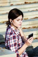 Image of beautiful stylish woman holding mobile phone. Young european girl standing at the street and using cellphone. Woman listening to music with the phone and having fun. Mobile internet concept. photo