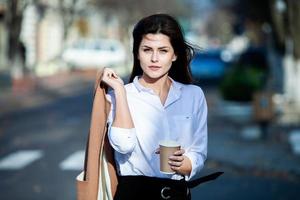 joven elegante bebiendo té en una calle de la ciudad. hipster chica europea con vaso de papel con leche. hermosa mujer joven con una taza de café en la calle de la ciudad. descanso. Café para llevar. foto