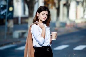 joven elegante bebiendo té en una calle de la ciudad. hipster chica europea con vaso de papel con leche. hermosa mujer joven con una taza de café en la calle de la ciudad. descanso. Café para llevar. foto