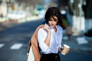 joven elegante bebiendo té en una calle de la ciudad. hipster chica europea con vaso de papel con leche. hermosa mujer joven con una taza de café en la calle de la ciudad. descanso. Café para llevar. foto