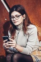 Image of beautiful stylish woman holding mobile phone. Young european girl standing at the street and using cellphone. Woman listening to music with the phone and having fun. Mobile internet concept. photo