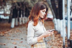 Image of beautiful stylish woman holding mobile phone. Young european girl standing at the street and using cellphone. Woman listening to music with the phone and having fun. Mobile internet concept. photo