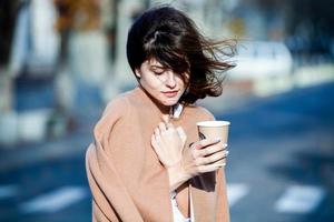 joven elegante bebiendo té en una calle de la ciudad. hipster chica europea con vaso de papel con leche. hermosa mujer joven con una taza de café en la calle de la ciudad. descanso. Café para llevar. foto