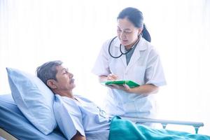 Asian woman professional doctor with notepad smiling, visiting, talking, and diagnosing the old man patient lying in patient bed at hospital ward photo