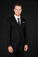 Portrait of young smart and handsome business man in black suit standing on isolated black background. photo