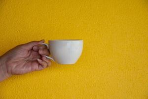 Young woman hand holding cup of hot coffee tea or water in  front of the isolated yellow texture  background photo