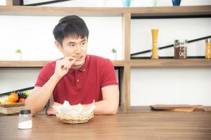 un joven asiático con una camiseta roja casual disfruta desayunando, comiendo papas fritas. joven cocinando comida en la cocina estilo loft foto