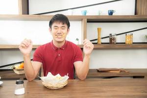 un joven asiático con una camiseta roja casual disfruta desayunando, comiendo papas fritas con risa. joven cocinando comida en la cocina estilo loft foto